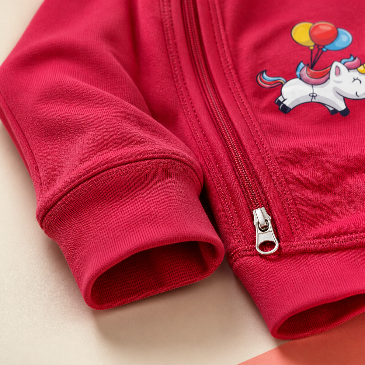 Kidz365 Pink Unicorn Balloons Footless Baby Sleepsuit - fabric detail closeup on deep fuchsia pink cotton showing left-chest placement graphic of kawaii chubby white baby unicorn with rainbow mane tail in pink blue red orange, golden horn, blissful closed-eye expression, floating with three-balloon cluster in yellow red and blue, lower two-way zip silver pull and inner zip guard visible